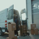 Courier loading cardboard boxes into a delivery van outside a warehouse.